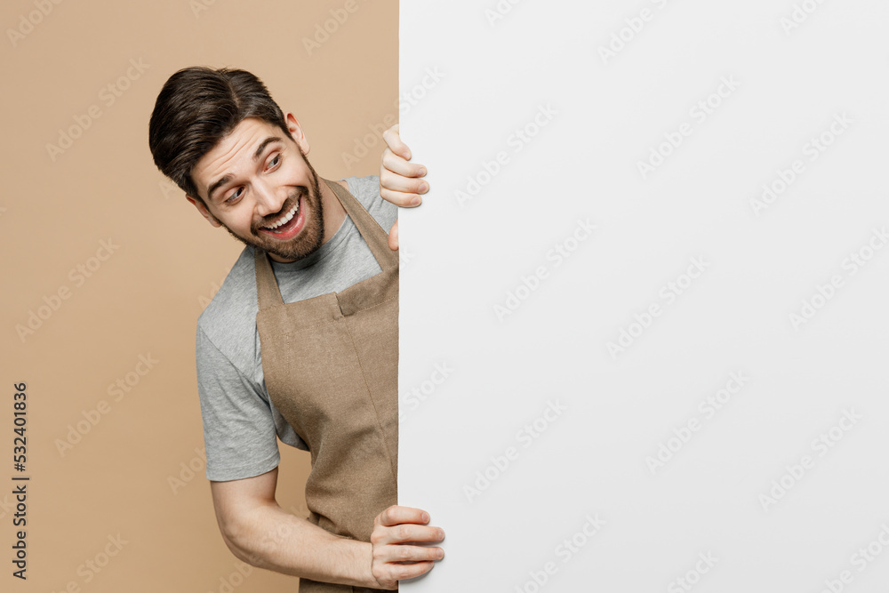 Young man barista barman employee in brown apron work in coffee shop look at big white billboard for content place text image isolated on plain pastel beige background Small business startup concept Young man barista barman employee in brown apron work in coffee shop look at big white billboard for content place text image isolated on plain pastel beige background Small business startup concept
