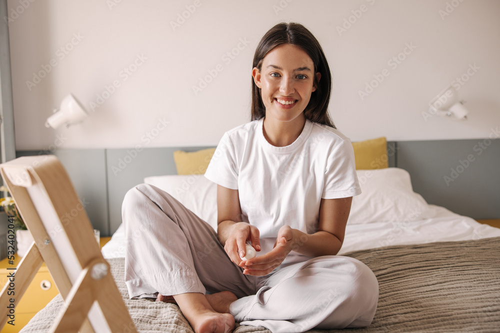 Cheerful young caucasian woman holding cream in her hands sits on bed in front of mirror. Brown hair girl wear pajamas smiling at camera. Concept emotions, beauty. Cheerful young caucasian woman holding cream in her hands sits on bed in front of mirror. Brown hair girl wear pajamas smiling at camera. Concept emotions, beauty.