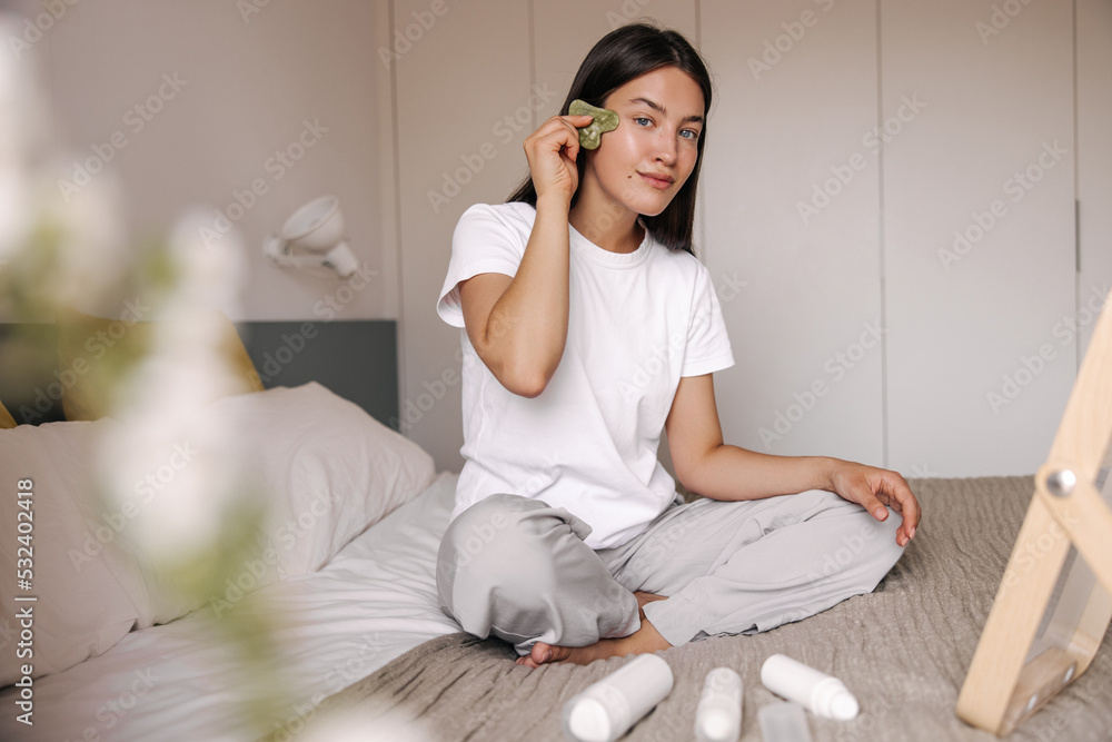 Pleasant young caucasian girl uses face massager sitting on bed at day. Brunette hair woman sitting in front of mirror. Concept beauty care, morning routine. Pleasant young caucasian girl uses face massager sitting on bed at day. Brunette hair woman sitting in front of mirror. Concept beauty care, morning routine.
