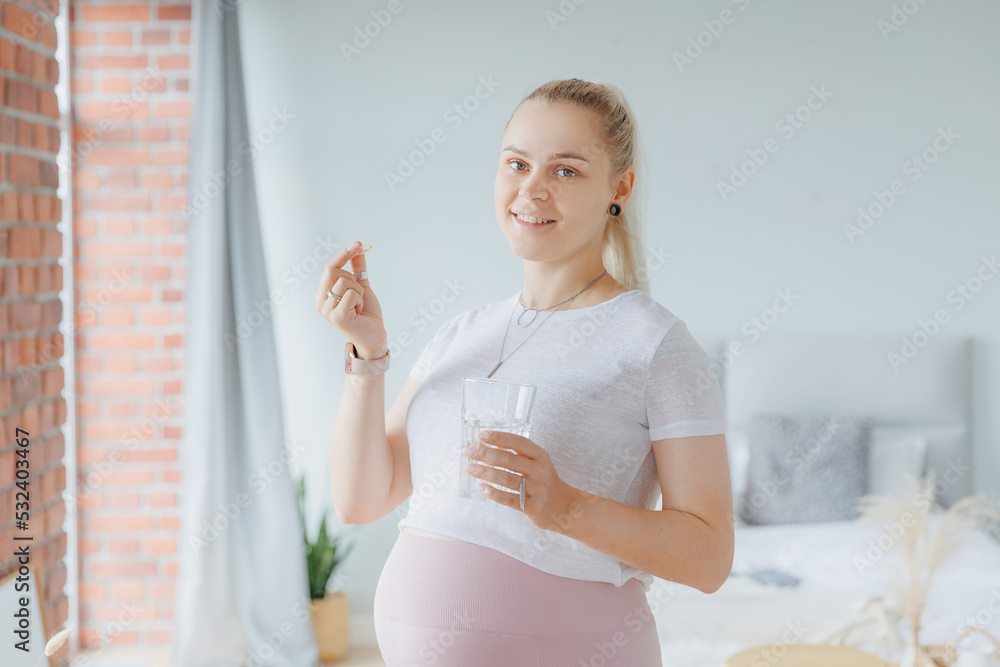 Happy young pregnant woman with white round pill and glass of water in hand, daily vitamins for hair, skin and health of baby Happy young pregnant woman with white round pill and glass of water in hand, daily vitamins for hair, skin and health of baby
