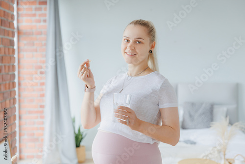 Happy young pregnant woman with white round pill and glass of water in hand, daily vitamins for hair, skin and health of baby