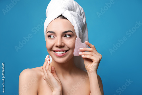 Beautiful young woman doing facial massage with gua sha tool on blue background