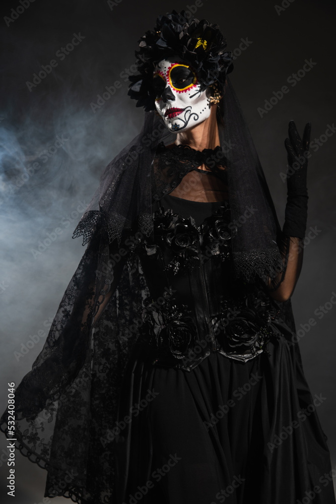 woman in black witch costume and creepy halloween makeup posing on dark background with fog.