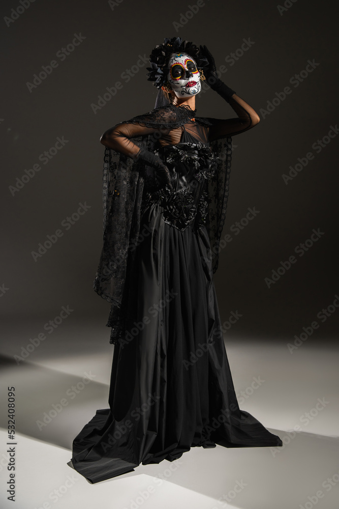 full length of woman with traditional mexican santa muerte makeup and costume posing with hand on waist on dark background.