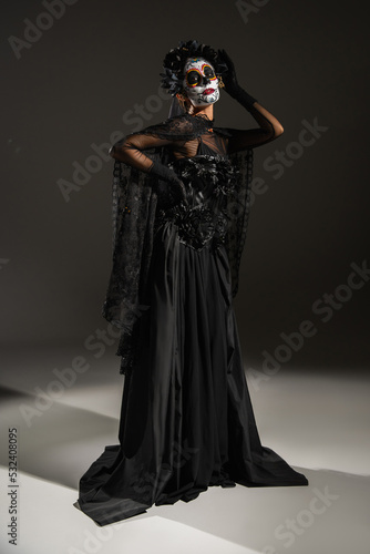 full length of woman with traditional mexican santa muerte makeup and costume posing with hand on waist on dark background.