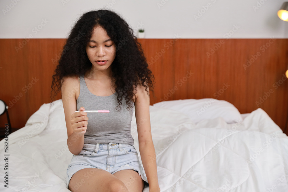 Shocked young woman looking at positive results of pregnancy test. Beautiful girl feeling sad and unexpected for bad news to be future parent on bed in bedroom. Single mon and expecting baby concept.