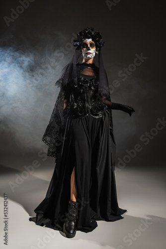 full length of woman in black halloween costume with veil and spooky makeup on dark background with smoke.