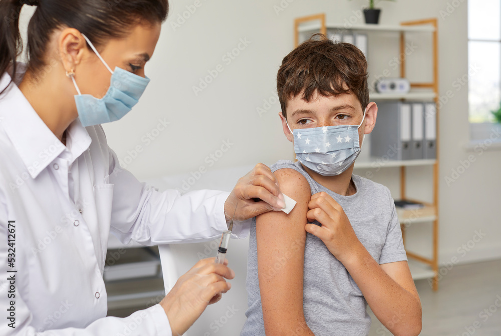 Doctor woman is giving a vaccine injection from flu, covid-19 to teen boy in shoulder in clinic. Healthcare, vaccination, immunization concept. Nurse and patient wearing protective medical masks.