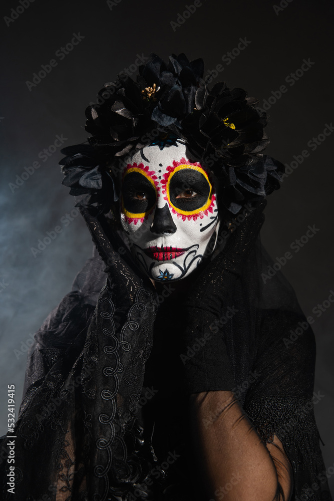 woman in black wreath holding hands near face with sugar skull makeup on dark smoky background.
