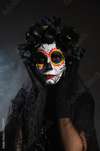 woman in black wreath holding hands near face with sugar skull makeup on dark smoky background.