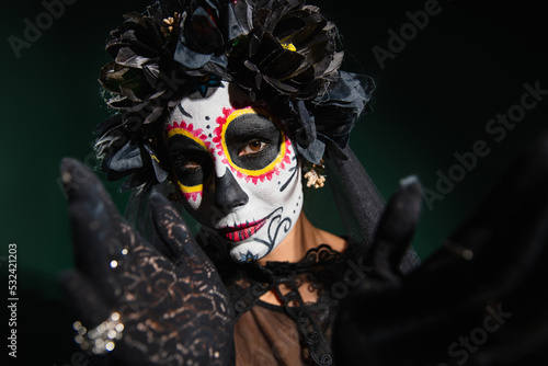 Woman in sugar skull makeup and wrench with black flowers looking at camera on dark green background.