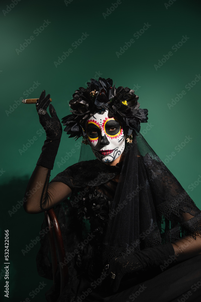 Woman in mexican santa muerte costume and makeup holding cigar and looking at camera on green background. Woman in mexican santa muerte costume and makeup holding cigar and looking at camera on green background.