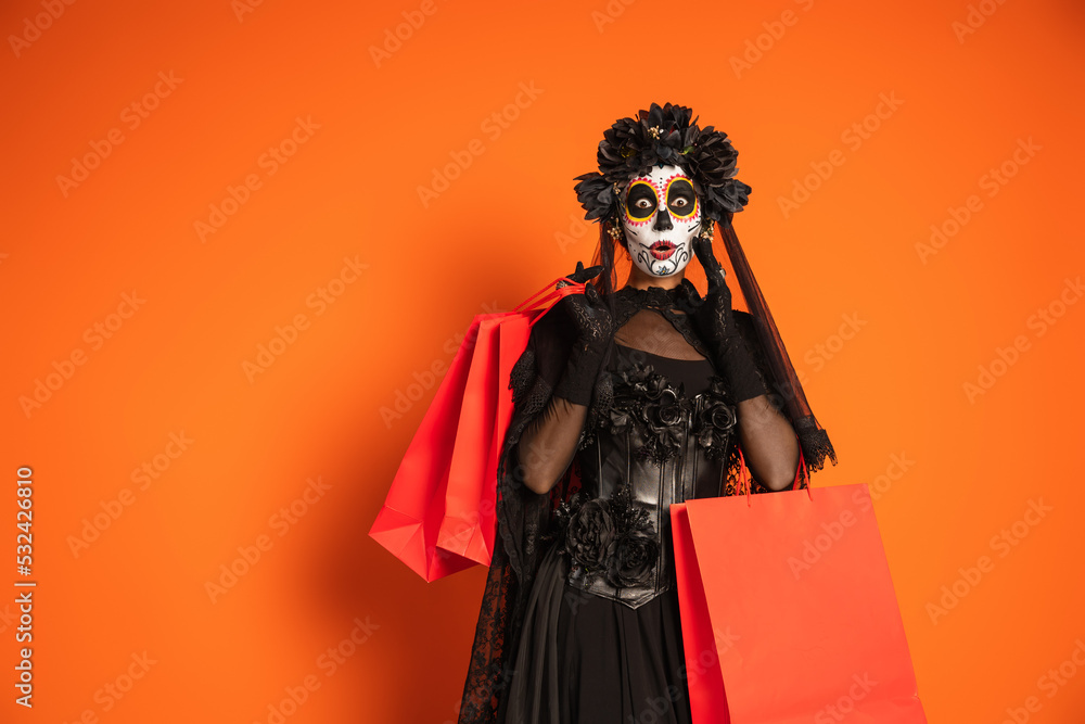 surprised woman in spooky halloween makeup and black costume holding shopping bags on orange background.