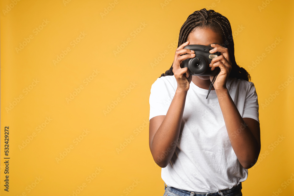 African-american woman taking photo of you. Female holding polaroid camera and pushing button standing isolated on yellow, making instant photo with retro camera African-american woman taking photo of you. Female holding polaroid camera and pushing button standing isolated on yellow, making instant photo with retro camera