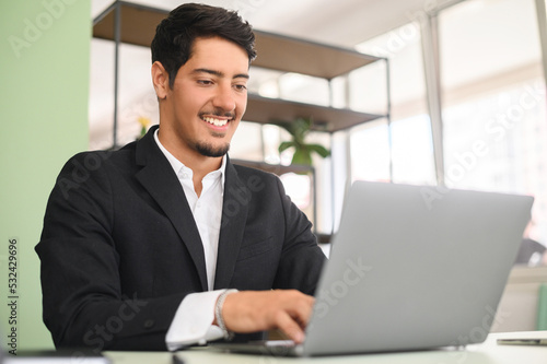 Smiling optimistic young latin entrepreneur wearing smart casual clothes, sitting at the desk, typing on keyboard, businessman professional is feeling ready to work and start new projects