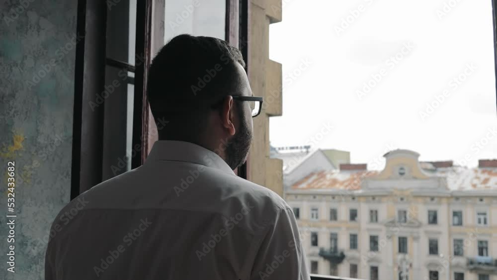 Rear view of a man approaching the window and putting on glasses looking through the window at the city Rear view of a man approaching the window and putting on glasses looking through the window at the city