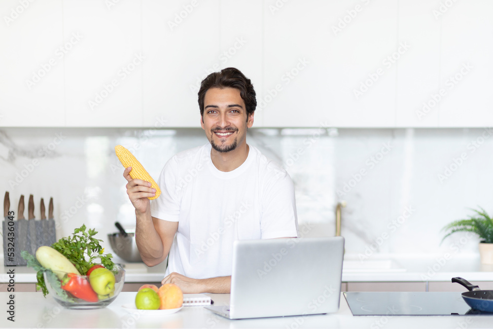 Smiling handsome man sitting in the kitchen and streaming on laptop, holding maize and explaining how to cook vegetables. Food blogger and healthy lifestyle concept Smiling handsome man sitting in the kitchen and streaming on laptop, holding maize and explaining how to cook vegetables. Food blogger and healthy lifestyle concept
