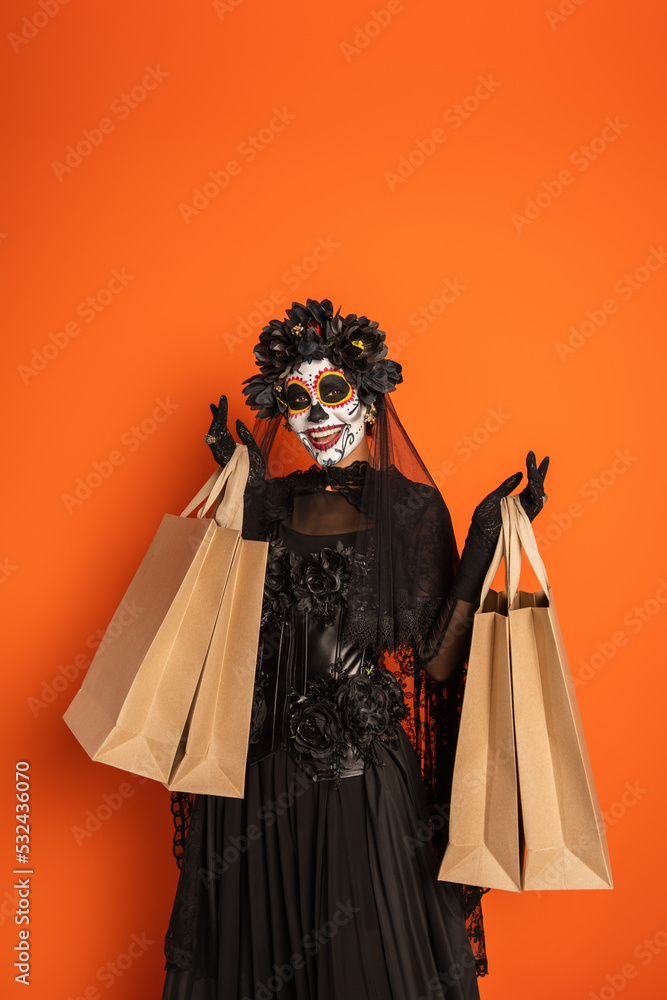 woman in mexican day of dead costume and catrina makeup smiling at camera with shopping bags isolated on orange.
