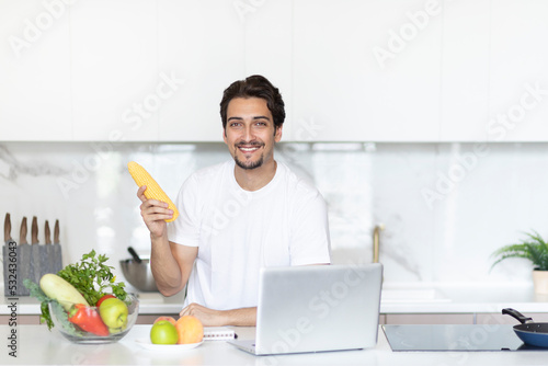Smiling handsome man sitting in the kitchen and streaming on laptop  holding maize and explaining how to cook vegetables. Food blogger and healthy lifestyle concept