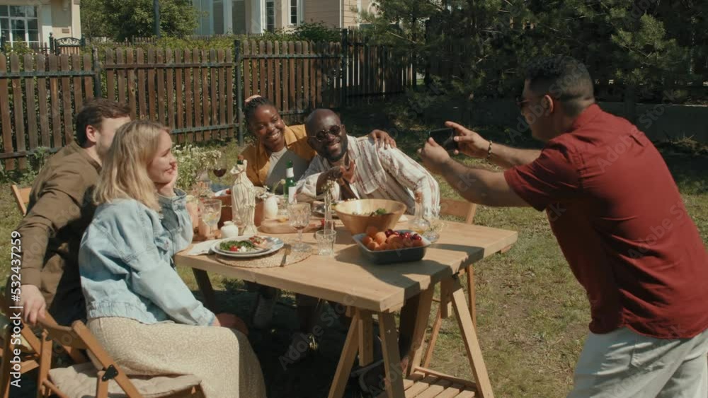 Middle Eastern man taking group photo of his friends sitting together at dining table outdoors on summer day using smartphone camera Middle Eastern man taking group photo of his friends sitting together at dining table outdoors on summer day using smartphone camera