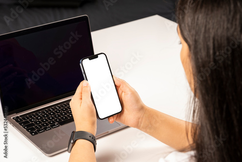 Fototapeta Naklejka Na Ścianę i Meble -  Smartphone mock up, young business woman holding smartphone mock up. Over shoulder view of caucasian lady sitting on office table using laptop and cell phone with white blank screen. Modern lifestyle.