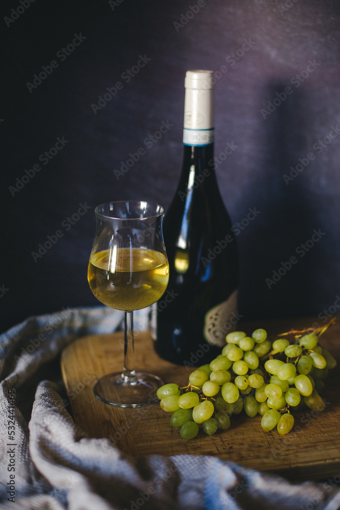 a glass of white wine on a wooden tray and a towel near a bottle of dry white wine and green fresh grapes side view on a black background romance all for one alcohol