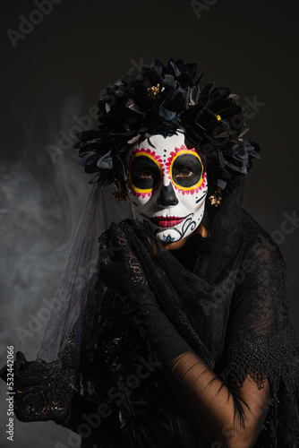 woman in black wreath and traditional santa muerte makeup looking at camera on dark foggy background.