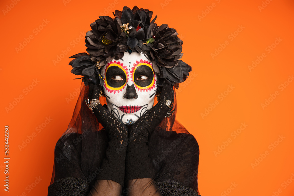 woman in traditional mexican day of dead makeup and black wreath holding hands near face and looking away isolated on orange.