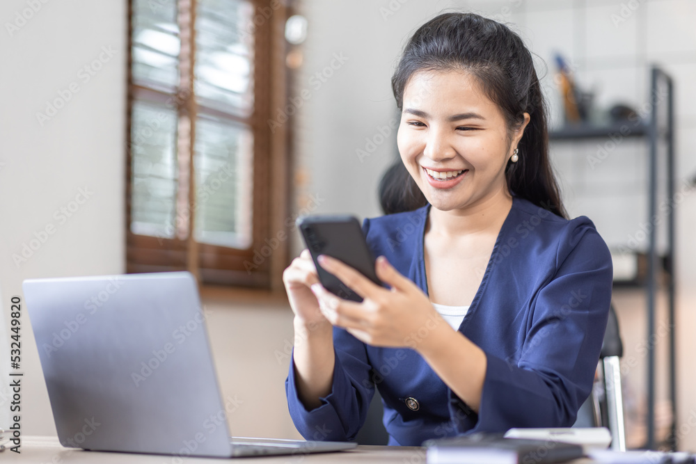 Happy Excited Asian young entrepreneur business woman using phone and laptop sitting on a desk officer in the day at workplace in office Happy Excited Asian young entrepreneur business woman using phone and laptop sitting on a desk officer in the day at workplace in office