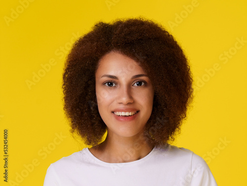 Portrait of african american woman with clean healthy skin and afro hair looking at camera smiling wearing white t-shirt isolated on yellow background. Young cheerful dark skin girl with curly hair