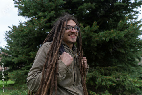 Young smiling boy with long dreadlocks posing next to trees. Traveler male wearing warm clothes posing in nature. Concept of scout, research, camping, travel and survival in nature.
