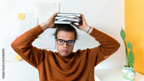 a young college student holding a pale of books on his head with a weary face, distance laerning concept photo