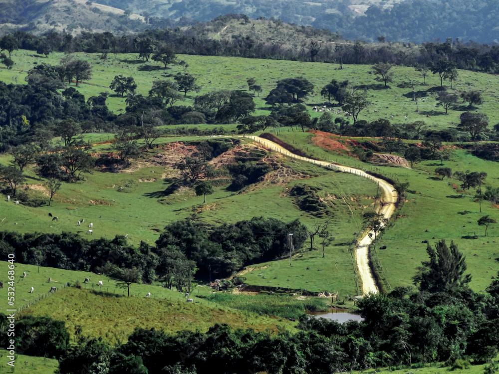 Estrada cortando fazenda, com lago artificial, muita vegetação e montanhas ao redor, localizada na região rural do bairro Jardim das Oliveiras, município de Esmeraldas, Minas Gerais, Brasil.