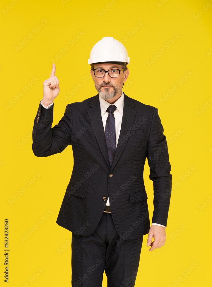 Idea, or pointing finger up middle aged businessman in black suit, construction helmet isolated on yellow background. Businessman in suit construction hard hat and eye glasses Idea, or pointing finger up middle aged businessman in black suit, construction helmet isolated on yellow background. Businessman in suit construction hard hat and eye glasses