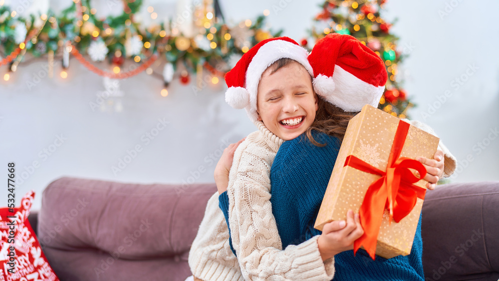 happy and loving mother and son, dressed in a Santa Claus hat, hug at home, sitting on the sofa against the background of a Christmas tree. child and woman are laughing happily while holding gift box. happy and loving mother and son, dressed in a Santa Claus hat, hug at home, sitting on the sofa against the background of a Christmas tree. child and woman are laughing happily while holding gift box.