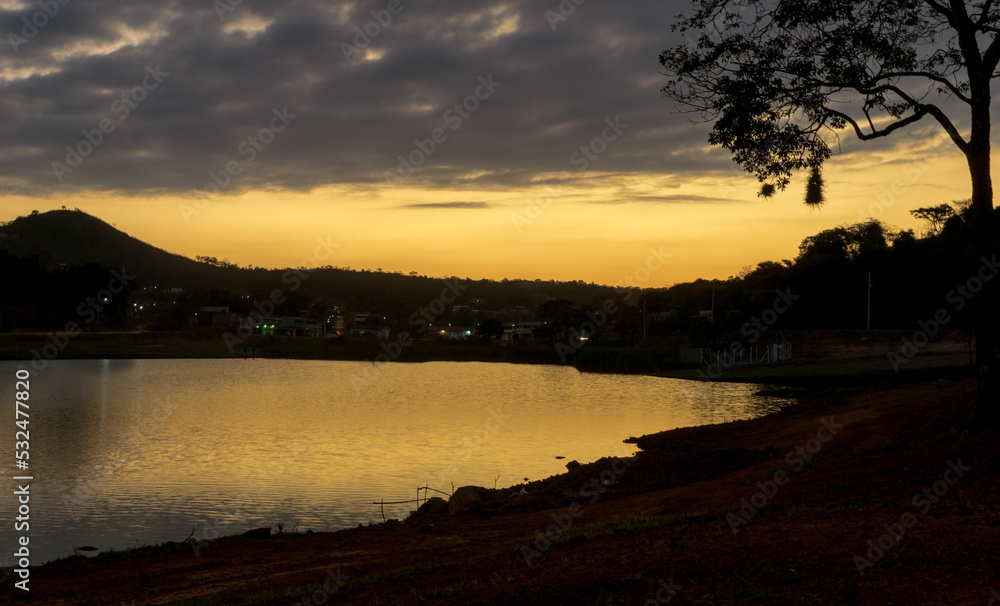 Pôr do sol entre a silueta de uma árvore ao entardecer com céu dourado e  limpo, com reflexo em lago ano bairro Jardim das Oliveiras, Esmeraldas, Minas Gerais.
