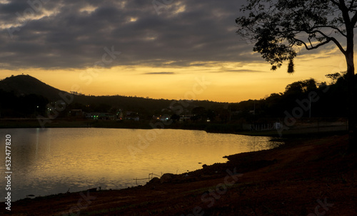 Pôr do sol entre a silueta de uma árvore ao entardecer com céu dourado e  limpo, com reflexo em lago ano bairro Jardim das Oliveiras, Esmeraldas, Minas Gerais.