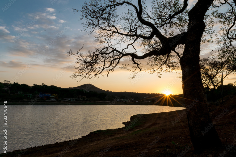 Pôr do sol entre nunvens e silueta de árvore ao entardecer com céu dourado e  limpo, com reflexo em lago no bairro Jardim das Oliveiras, Esmeraldas, Minas Gerais.