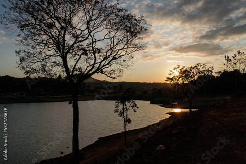 Lindo p  r do sol entre nunvens e silueta de   rvore ao entardecer com c  u dourado e  limpo  com reflexo em lagoa no bairro Jardim das Oliveiras  Esmeraldas  Minas Gerais.