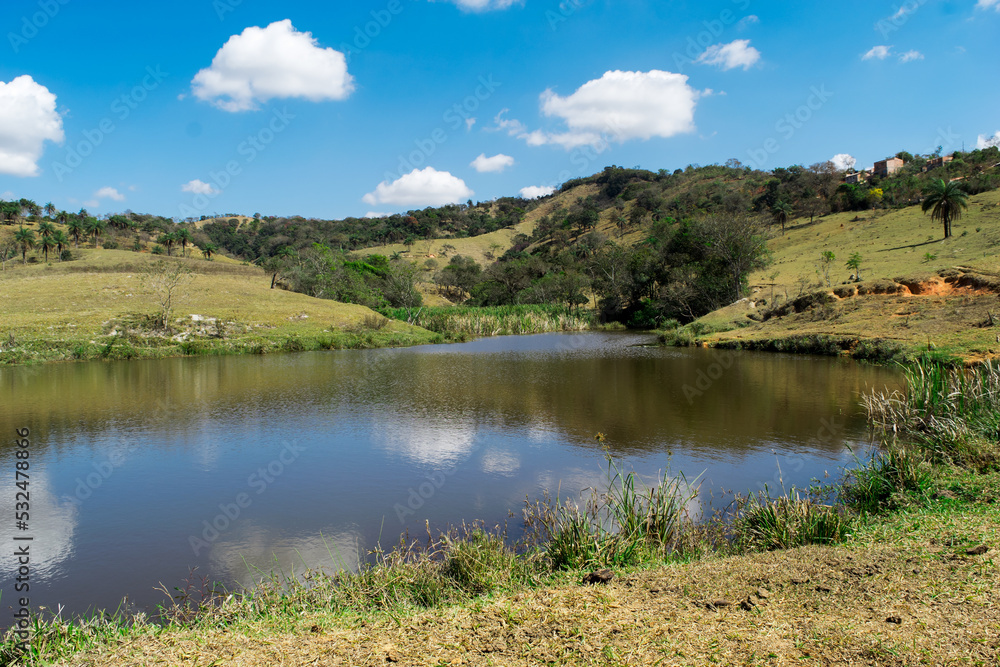 Paisagem de verão em fazenda, com muita vegetação ao redor, lindo lago, céu azul tudo em meio a montanhas, no bairro Jardim das Oliveiras, Esmeraldas, Minas Gerais, Brasil.