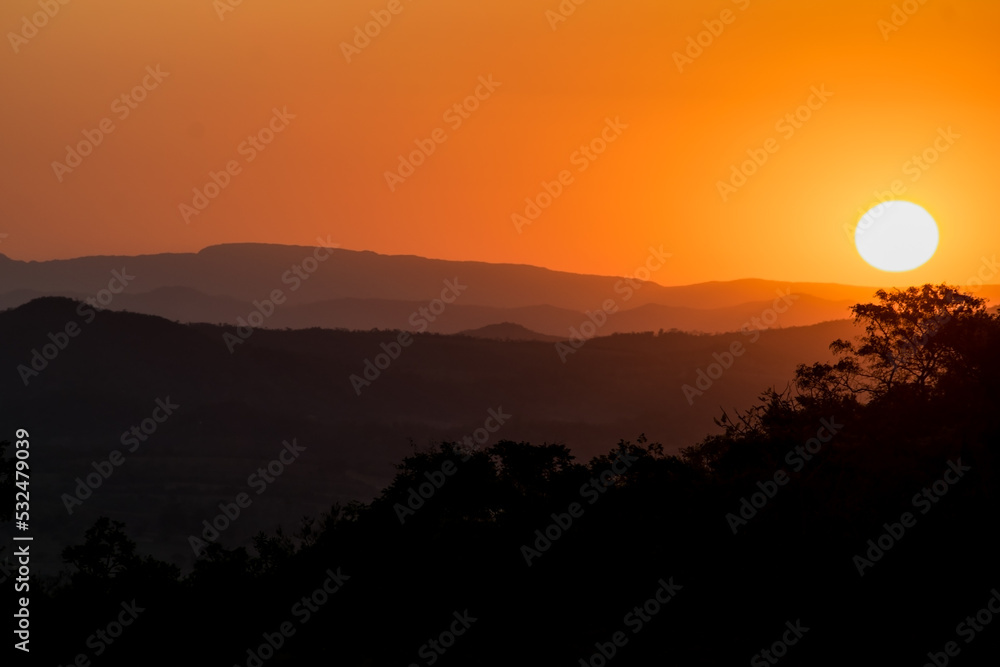 Pôr do sol com nuvens e silhueta de árvores ao entardecer, com céu dourado e limpo, de cima de montanha no bairro Jardim das Oliveiras, Esmeraldas, Minas Gerais, Brasil