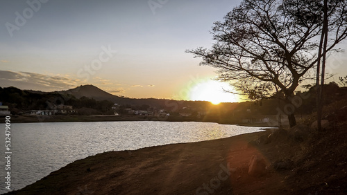 Pôr do sol entre a silueta de uma árvore ao entardecer com céu dourado e limpo, com reflexo em lago a no bairro Jardim das Oliveiras, Esmeraldas, Minas Gerais, Brasil