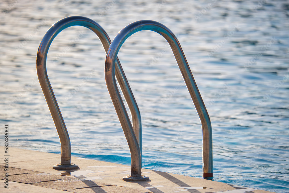 Close up of swimming pool stainless steel handrail descending into tortoise clear pool water. Accessibility of recreational activities concept Close up of swimming pool stainless steel handrail descending into tortoise clear pool water. Accessibility of recreational activities concept