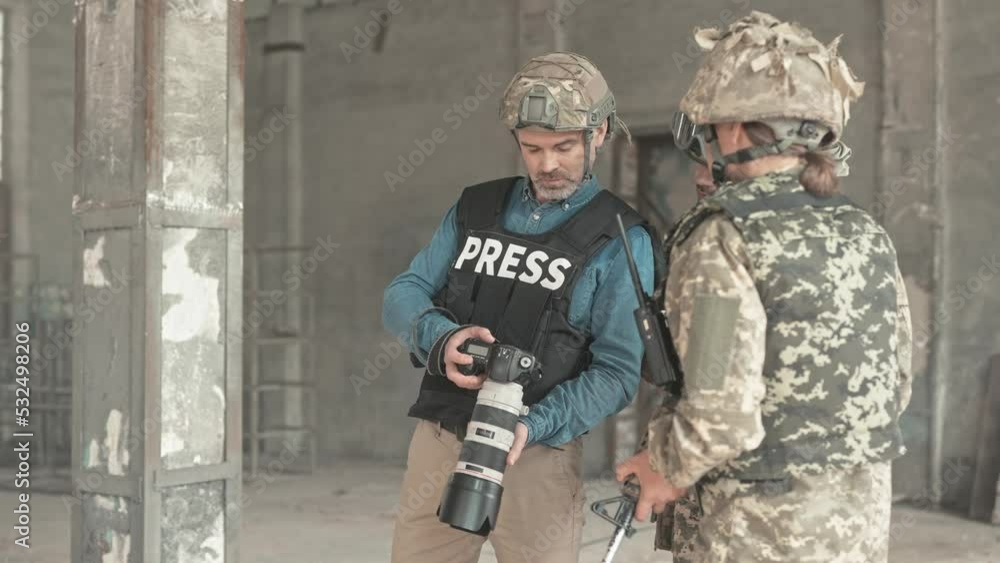 Close up of middle-aged professional photographer journalist working at war zone showing photos to soldiers in camouflage uniform with rifles in old ruined building hiding from bomb shelling Close up of middle-aged professional photographer journalist working at war zone showing photos to soldiers in camouflage uniform with rifles in old ruined building hiding from bomb shelling