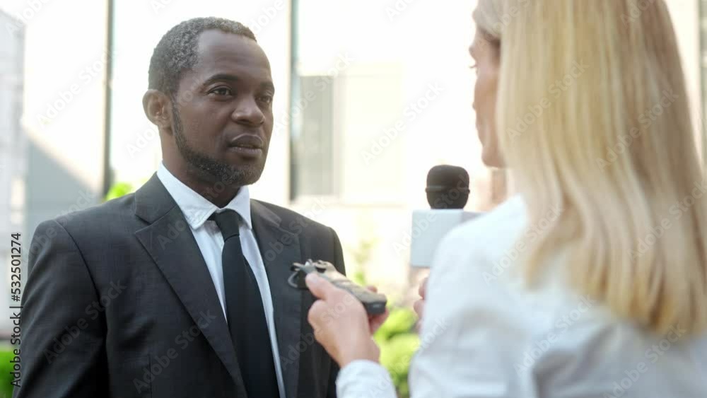 Close up of African American handsome serious politician in black suit giving comments to young Caucasian female journalist. Businessman giving interview to woman reporter in the street. News concept Close up of African American handsome serious politician in black suit giving comments to young Caucasian female journalist. Businessman giving interview to woman reporter in the street. News concept