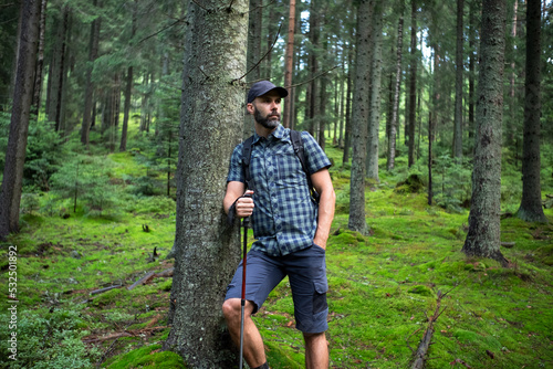 Hiker beard man in a green mountain forest. Dressed up in a cap, shorts, and plaid shirt with a backpack and trekking sticks