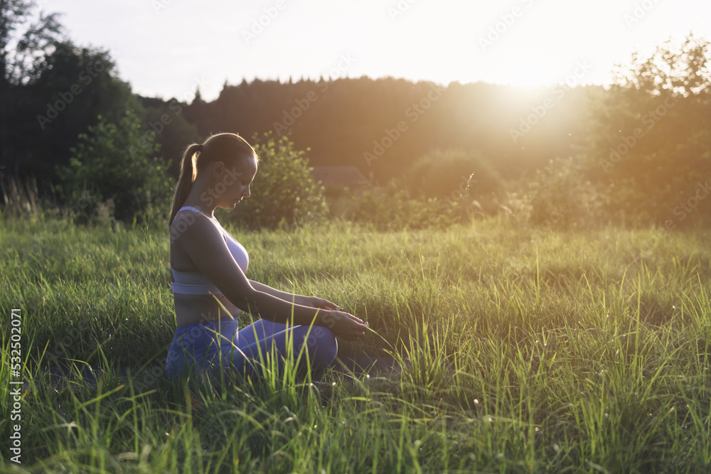 a girl of light appearance at sunset in a park doing exercises in sportswear, sitting on the grass, a silhouette of a woman, there is a place for an inscription