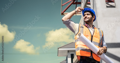 Portrait of multiethnic architect with blueprints in contruction site. Mature middle eastern contractor holding roll of architectural projects while looking at Sky. Happy latin man in safety wear.