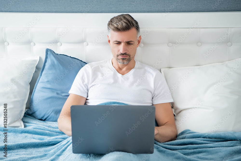 concentrated mature man working on laptop in bed concentrated mature man working on laptop in bed