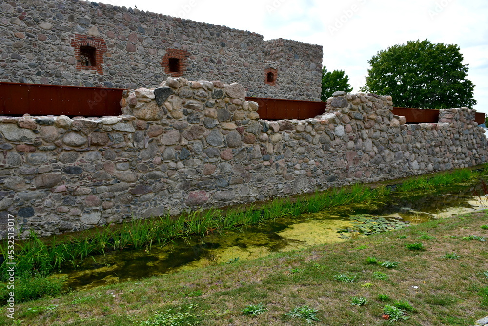 A close up on a moat and external wall of a replica of a medieval castle made out of stones, boulders, and rocks, seen next to a vast public park, walkway, and square full of flowers and other flora